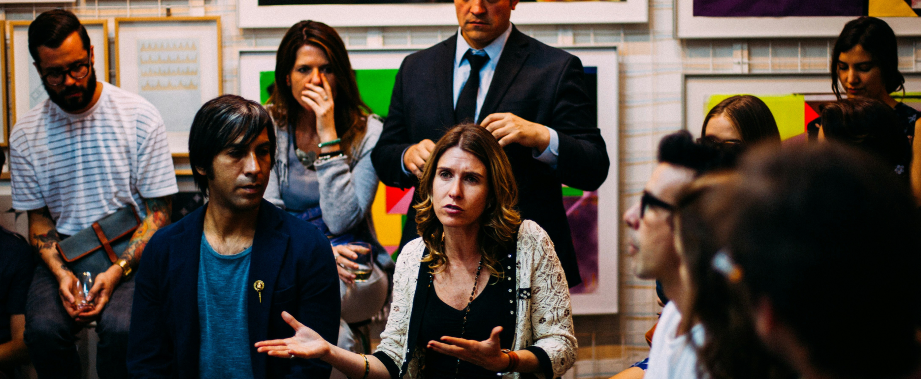 Image shows a town hall meeting in which a woman is speaking to around 30 people. This image is representative of a community benefits event.