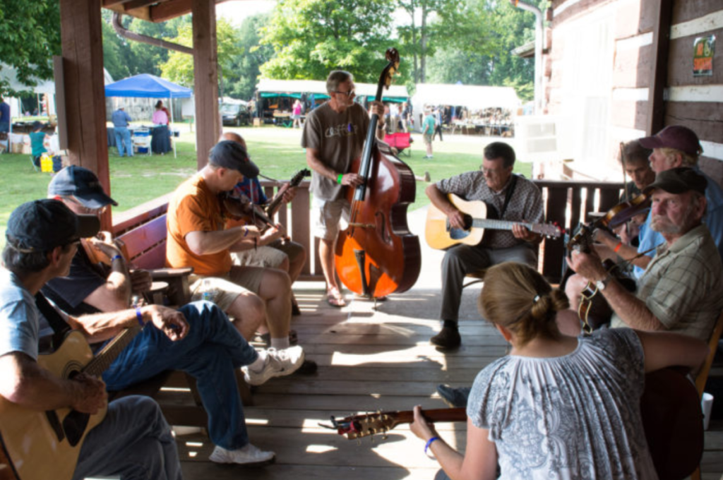 Image shows a folk band of 10 musicians sitting on a porch playing old time music.