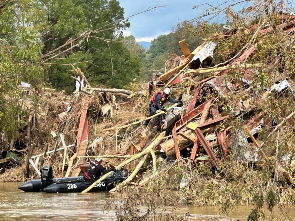 Image shows a first responder climbing along a pile of wreckage caused by hurricane Helene. The pile comprises fallen trees and destroyed scaffolding and showcases the impacts of flood risks.